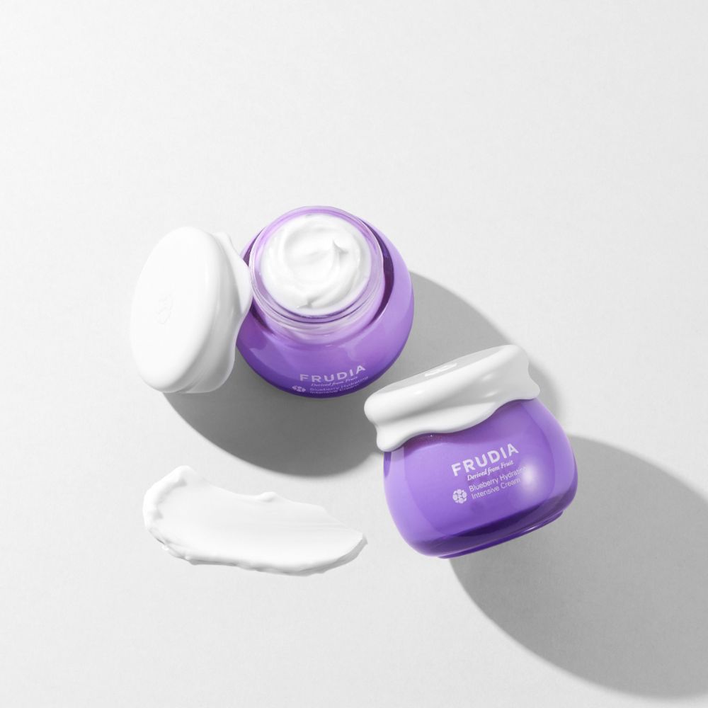 Frudia Blueberry Hydrating Intensive Cream face moisturizer in a jar with a blue lid and fresh blueberries beside it.