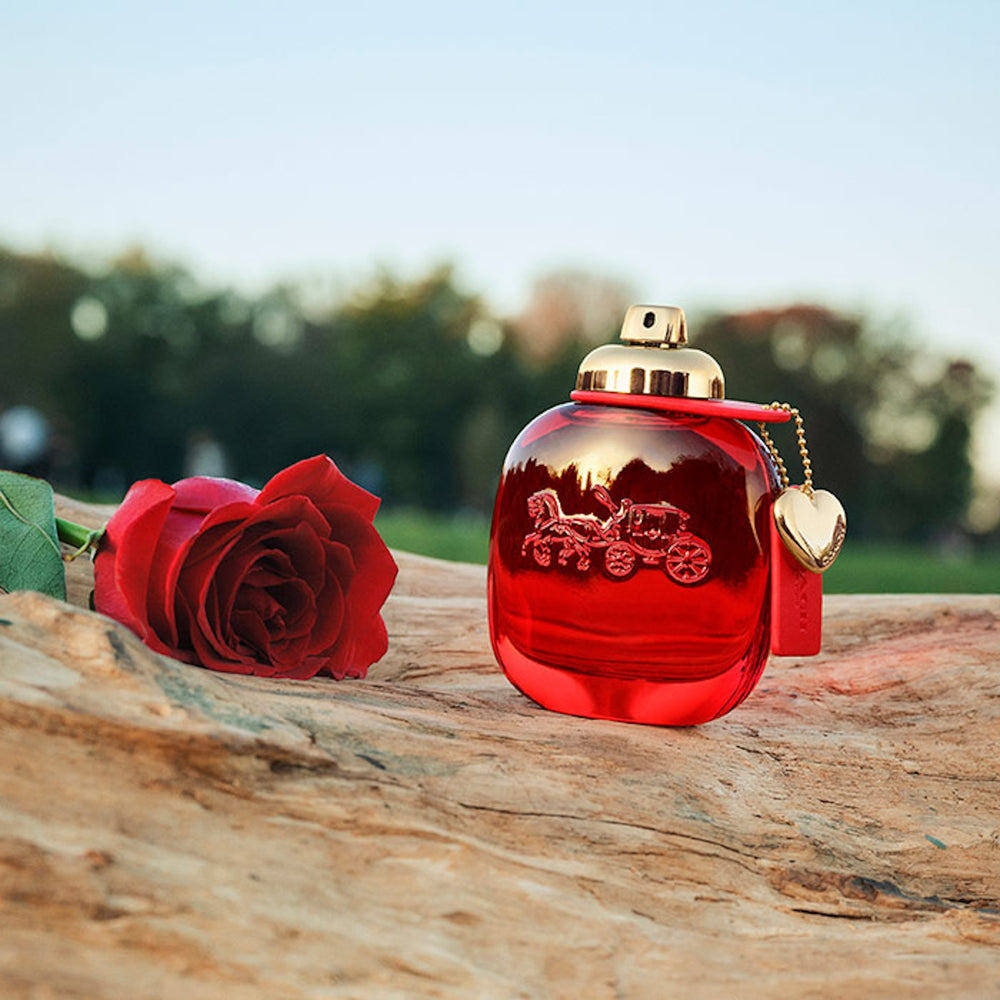 Red bottle of Coach Love perfume displayed on wooden platform with a red rose next to the perfume. 