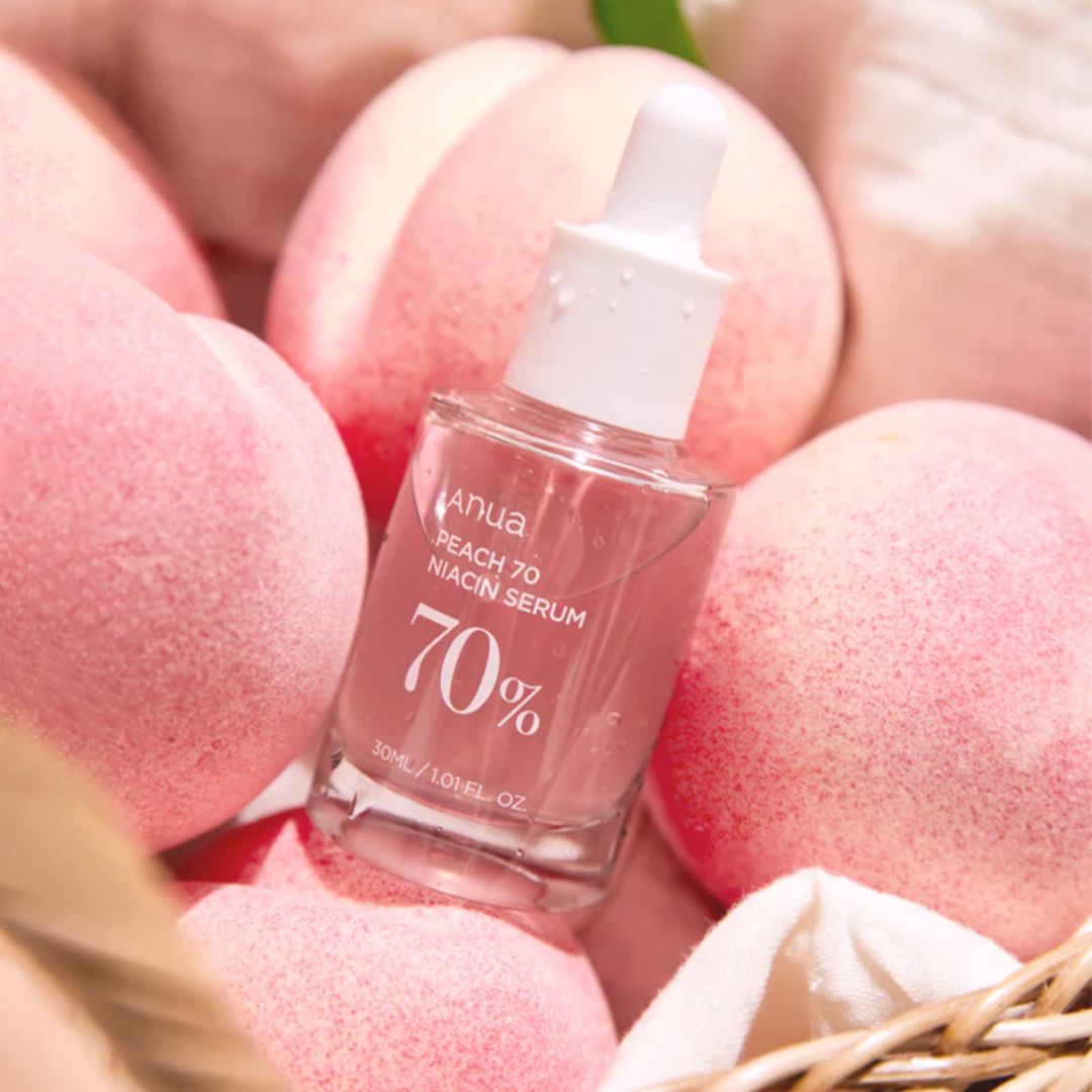 Anua Peach 70 Niacinamide Serum 30ml bottle with a peach-colored label and dropper cap, next to fresh peaches.